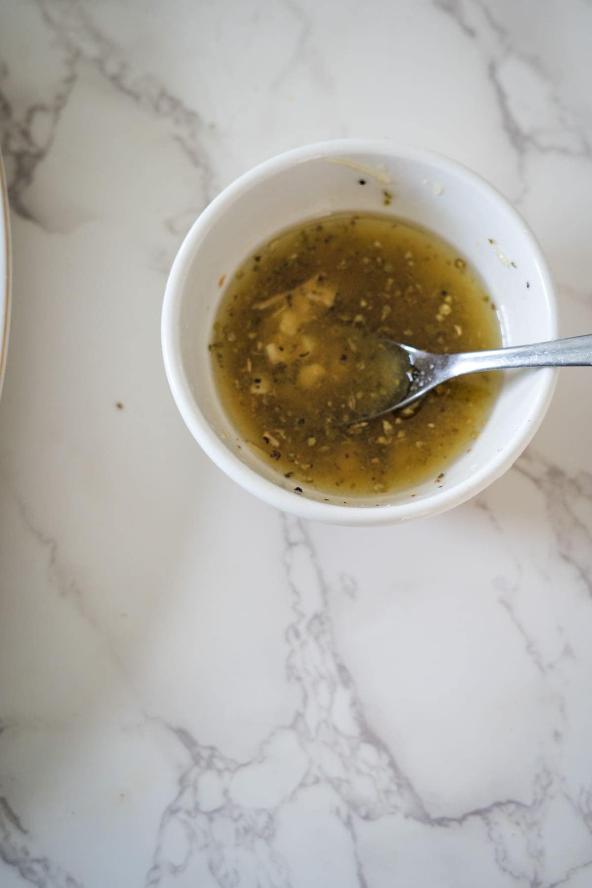 A round white bowl containing a light brown liquid with tiny chopped ingredients, reminiscent of a Greek village salad, and a spoon inside, resting on a white marbled surface.