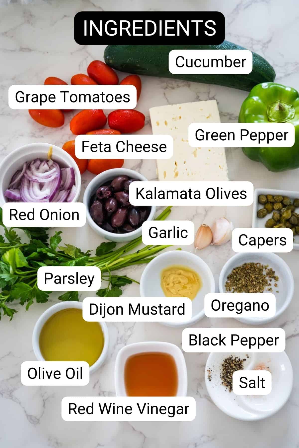 Ingredients for a dish displayed on a marble surface, including cucumber, grape tomatoes, green pepper, feta cheese, red onion, Kalamata olives, parsley, garlic, capers, oregano, black pepper, salt, Dijon mustard, olive oil, and red wine vinegar.