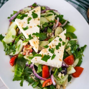 A fresh salad featuring cucumber, tomatoes, red onion, bell peppers, olives, and topped with two triangular pieces of white cheese, garnished with chopped parsley.