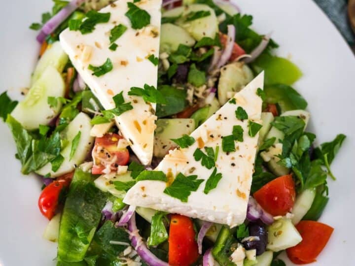 A fresh salad featuring cucumber, tomatoes, red onion, bell peppers, olives, and topped with two triangular pieces of white cheese, garnished with chopped parsley.