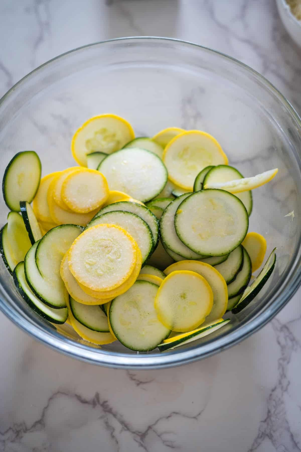 A glass bowl containing sliced yellow and green zucchini placed on a marble countertop.