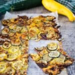 A baked zucchini and yellow squash flatbread on parchment paper, with a piece cut out. Fresh zucchini and yellow squash are placed above the flatbread on a gray surface.