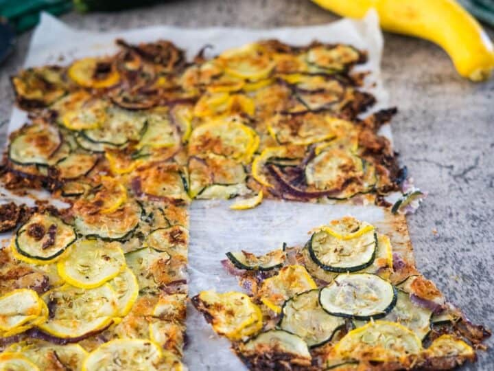 A baked zucchini and yellow squash flatbread on parchment paper, with a piece cut out. Fresh zucchini and yellow squash are placed above the flatbread on a gray surface.