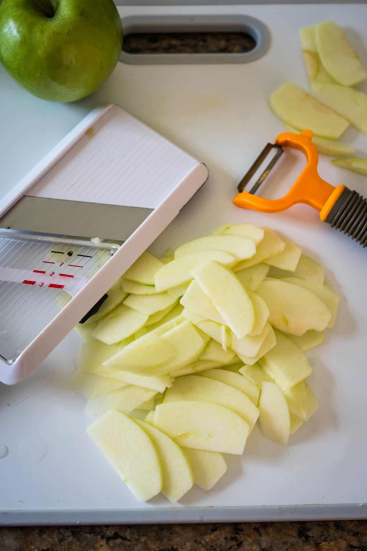 A green apple, a pile of thinly sliced apple pieces, a mandoline slicer, and an orange peeler rest on a white cutting board.