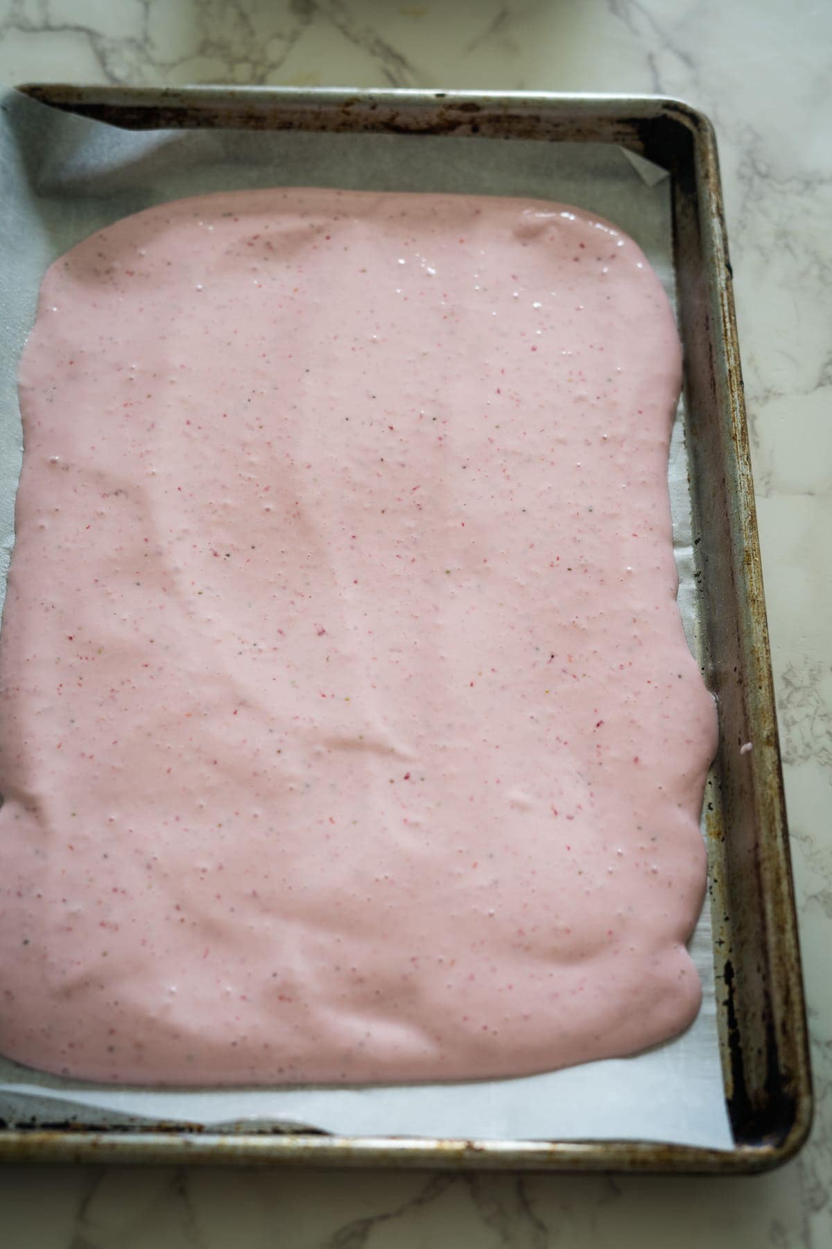 A baking tray with a layer of pink batter spread evenly on parchment paper, reminiscent of preparing frozen cottage cheese bark.