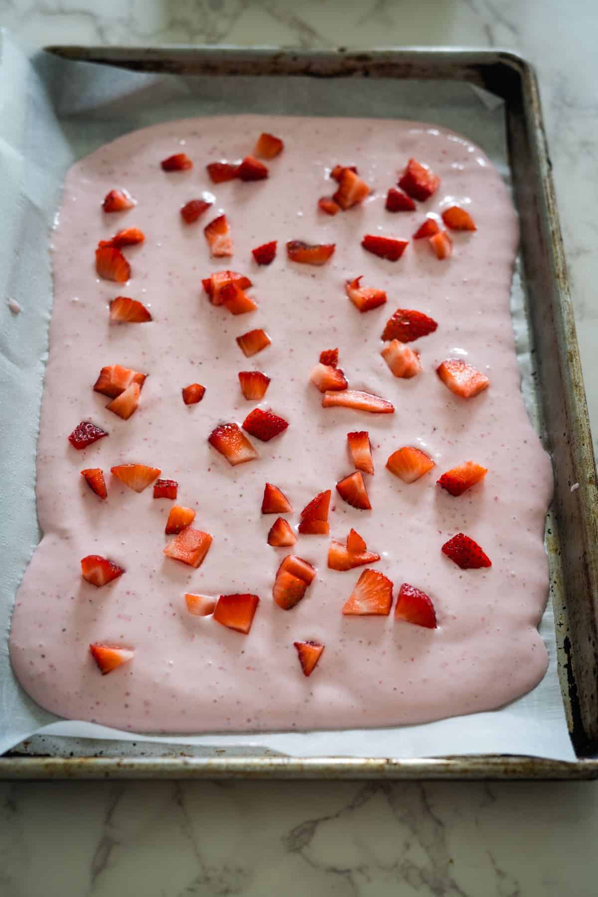 A baking tray on a marble countertop contains a pink yogurt mixture topped with chopped strawberries, resembling a delightful frozen cottage cheese bark.