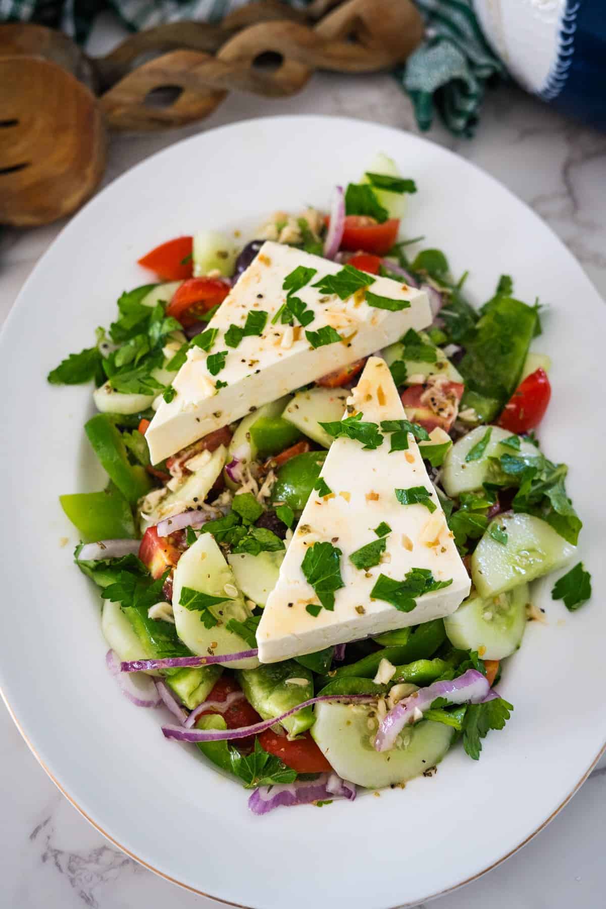 A white plate filled with a fresh Greek village salad featuring cucumber, tomato, red onion, green pepper, herbs, and two large wedges of cheese on top.