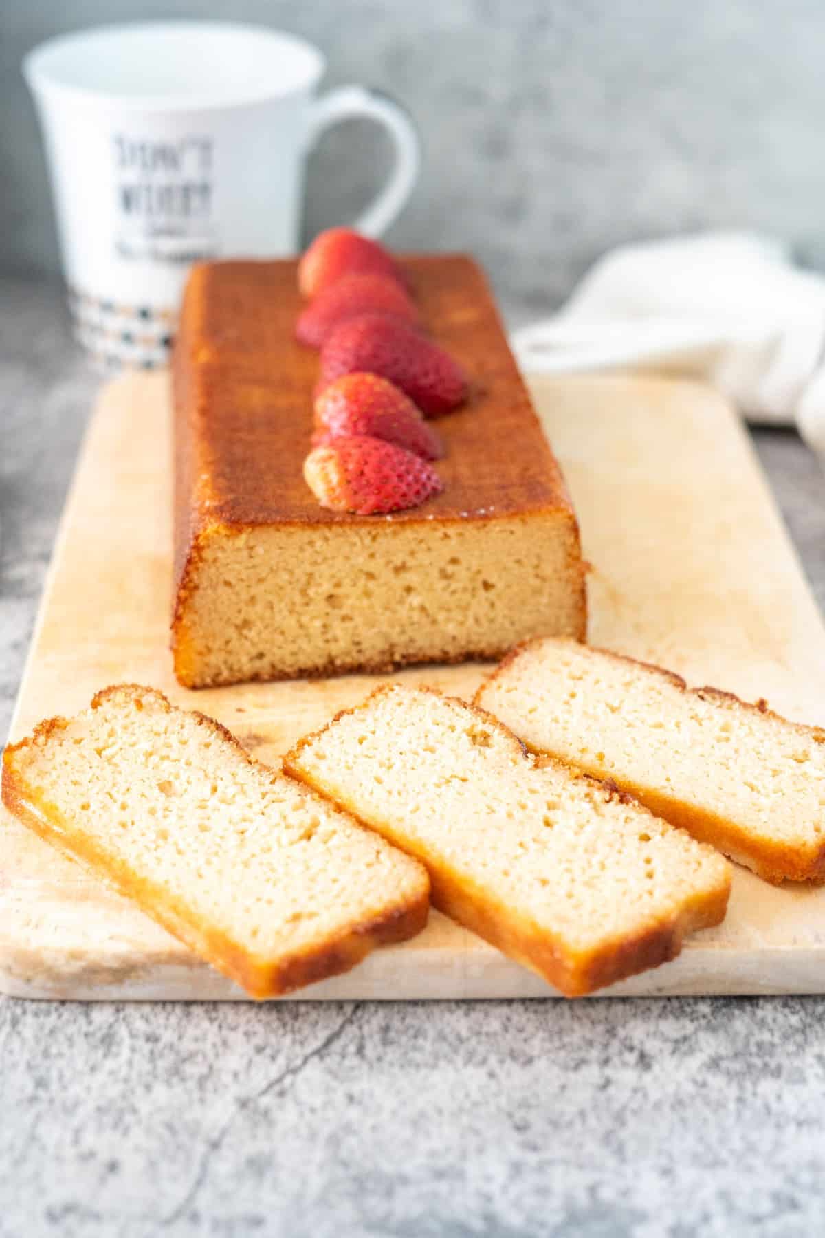 A loaf of pound cake topped with strawberries on a wooden cutting board with three slices in front. A white cup is in the background.