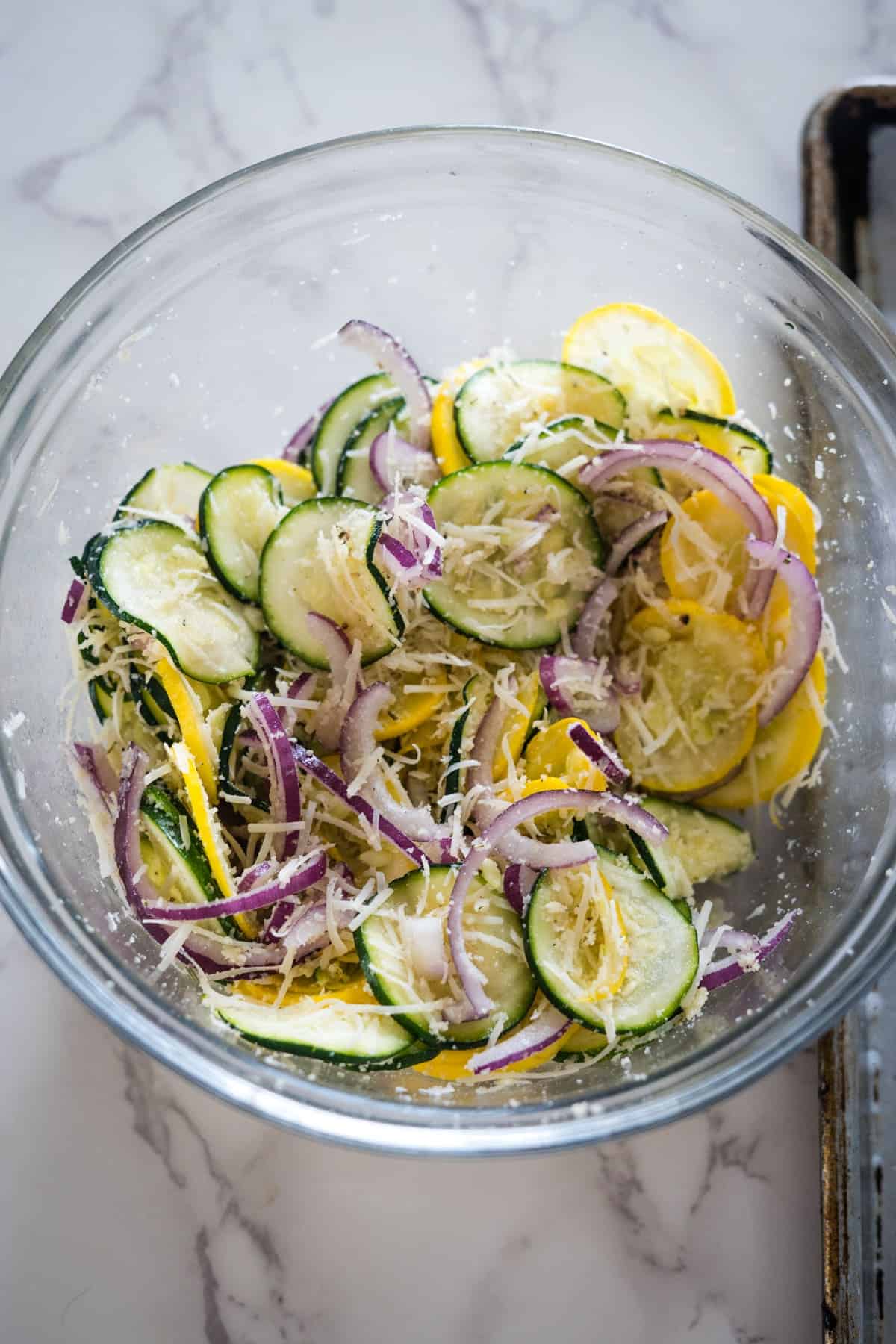 A glass bowl filled with sliced zucchini, yellow squash, red onion, and grated cheese, all mixed together.