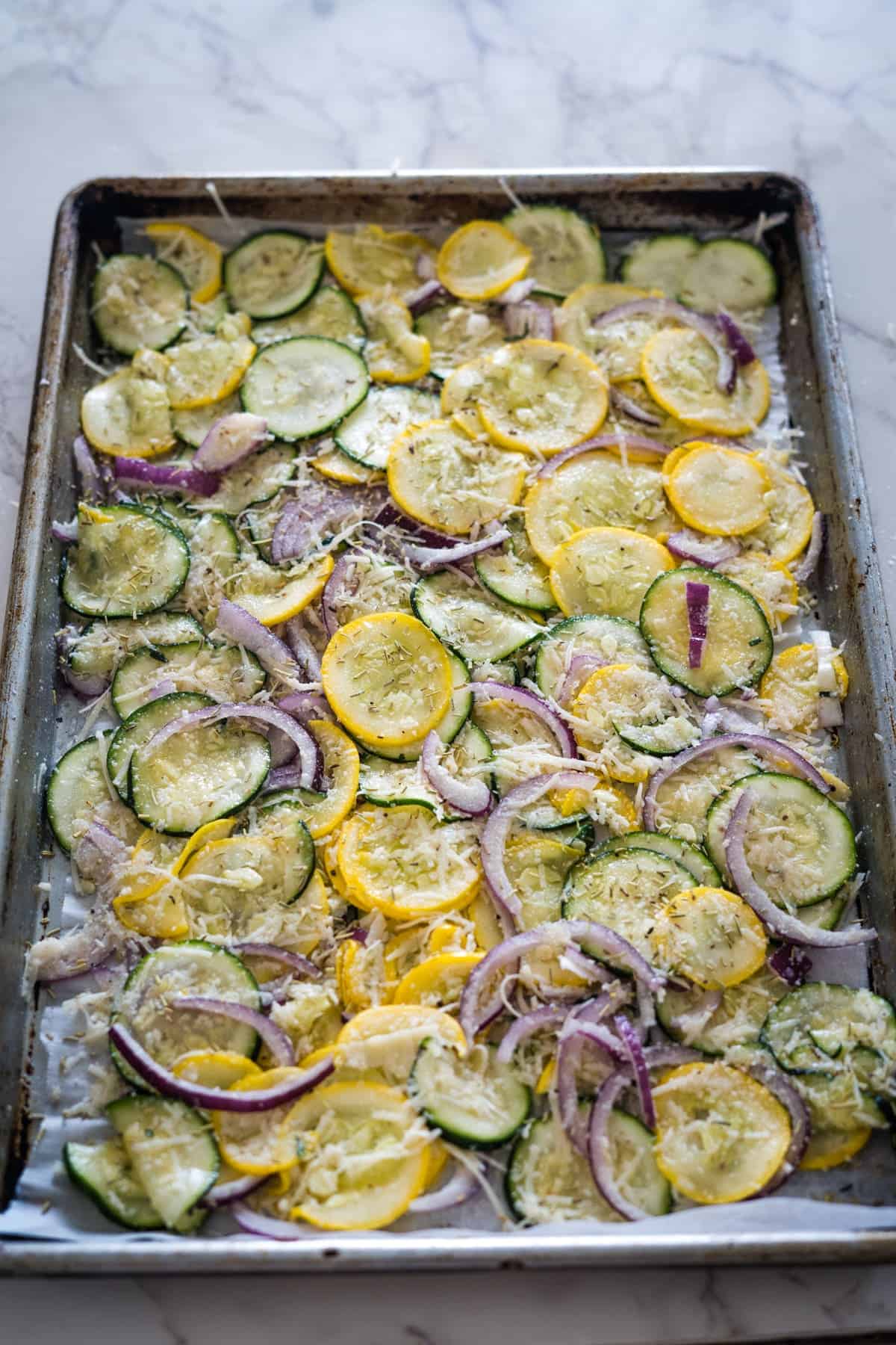 A baking sheet with sliced yellow squash, zucchini, and red onions, all seasoned and ready to be roasted.