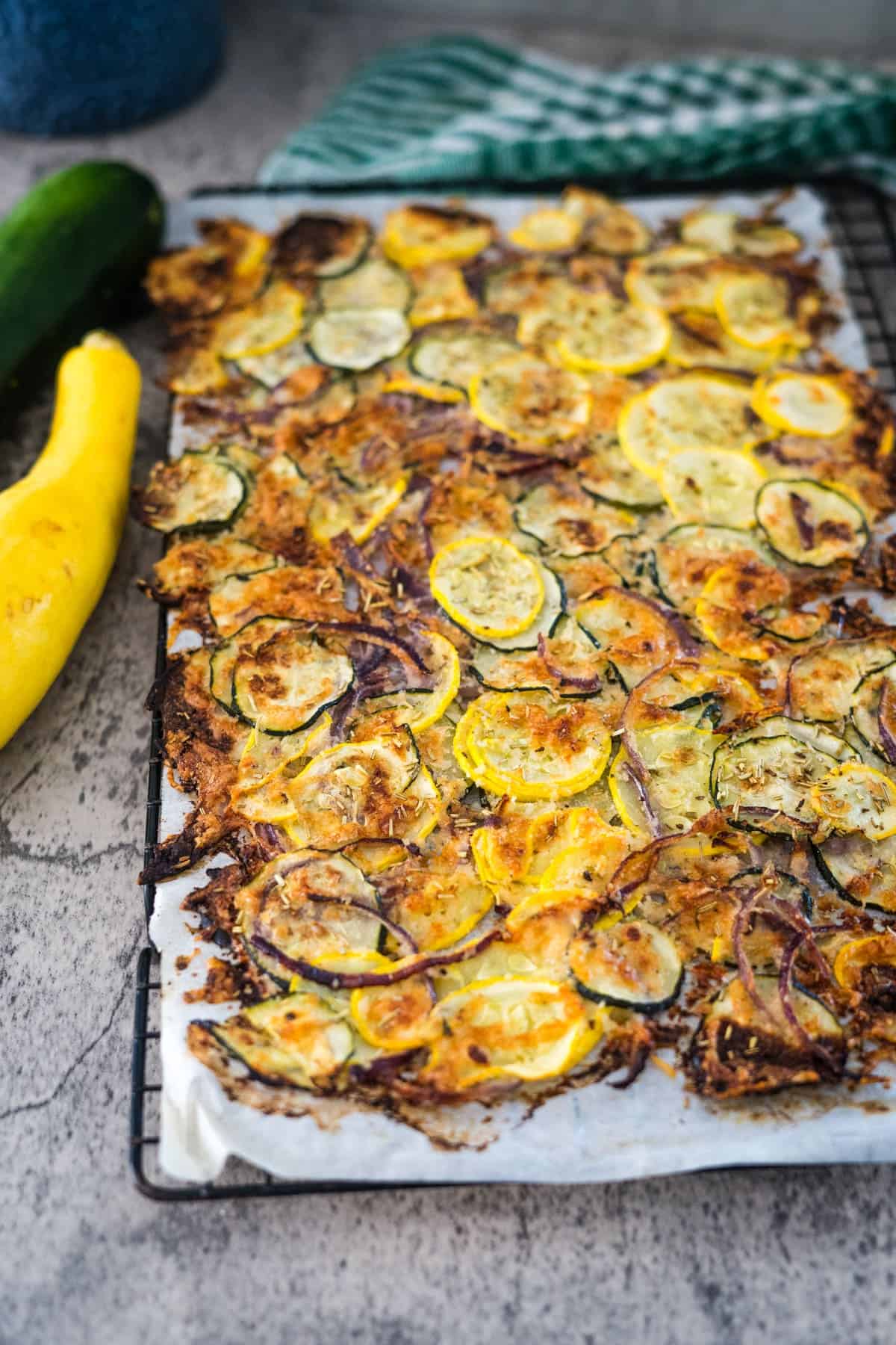 A sheet pan topped with baked zucchini and yellow squash slices mixed with red onions, grated cheese, and herbs. Two fresh squashes are placed on the side.