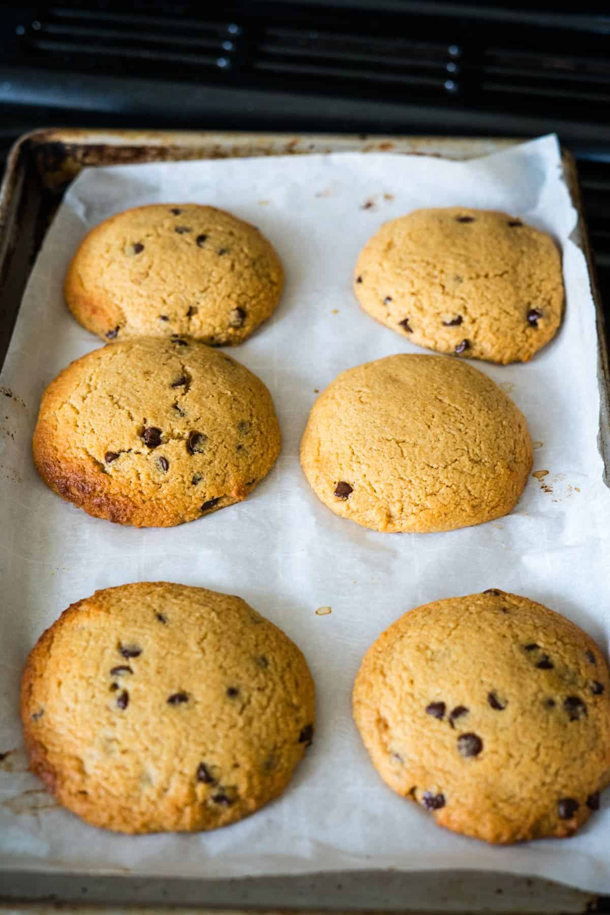 A baking tray with six freshly baked round cookies, some with visible chocolate chips, placed on parchment paper.