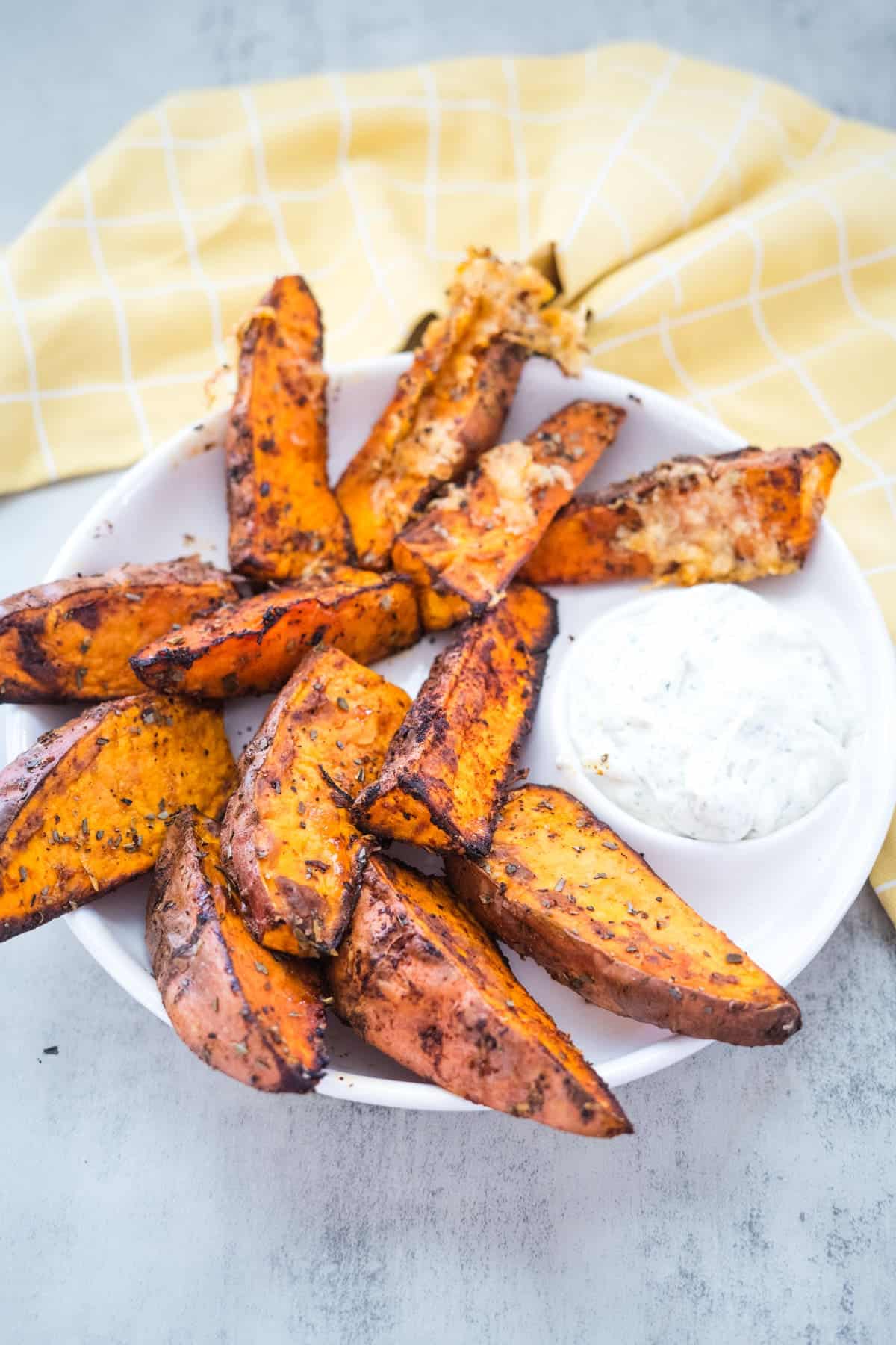 A plate of seasoned potato wedges with a side of white dipping sauce, placed on a white surface with a yellow checkered cloth in the background.