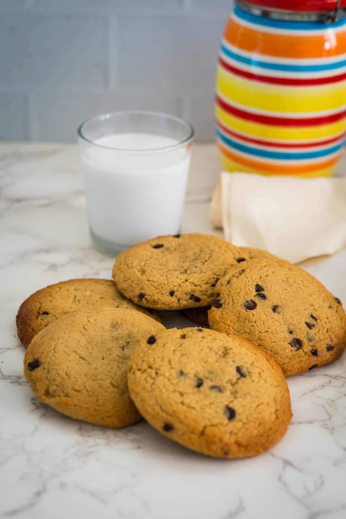 A stack of chocolate chip cookies is placed on a marble countertop beside a glass of milk and a colorful striped vase.