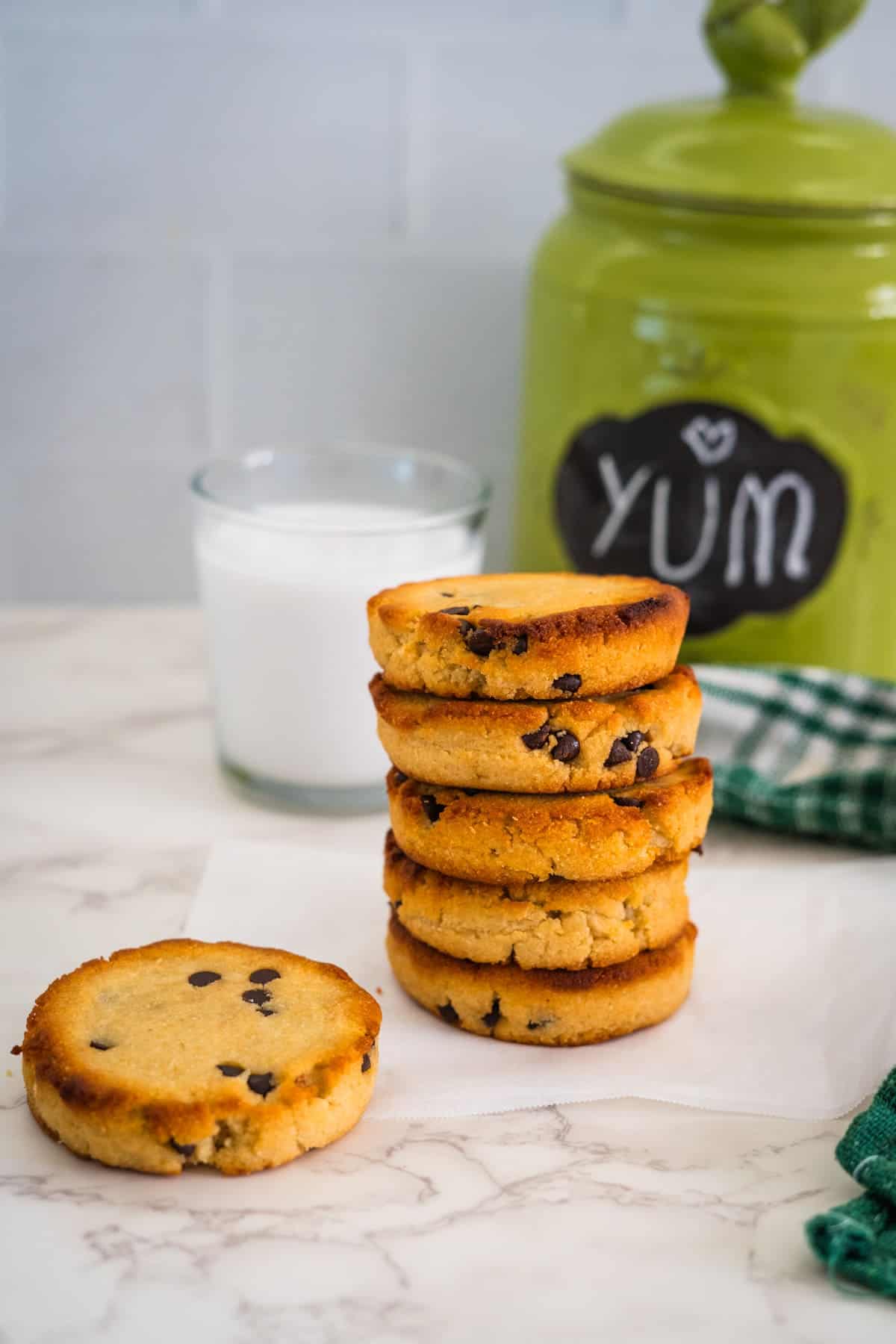 A stack of chocolate chip cookies on a white surface next to a glass of milk and a green jar with "Yum" written on it, perfect for pairing with a keto ice cream sandwich.