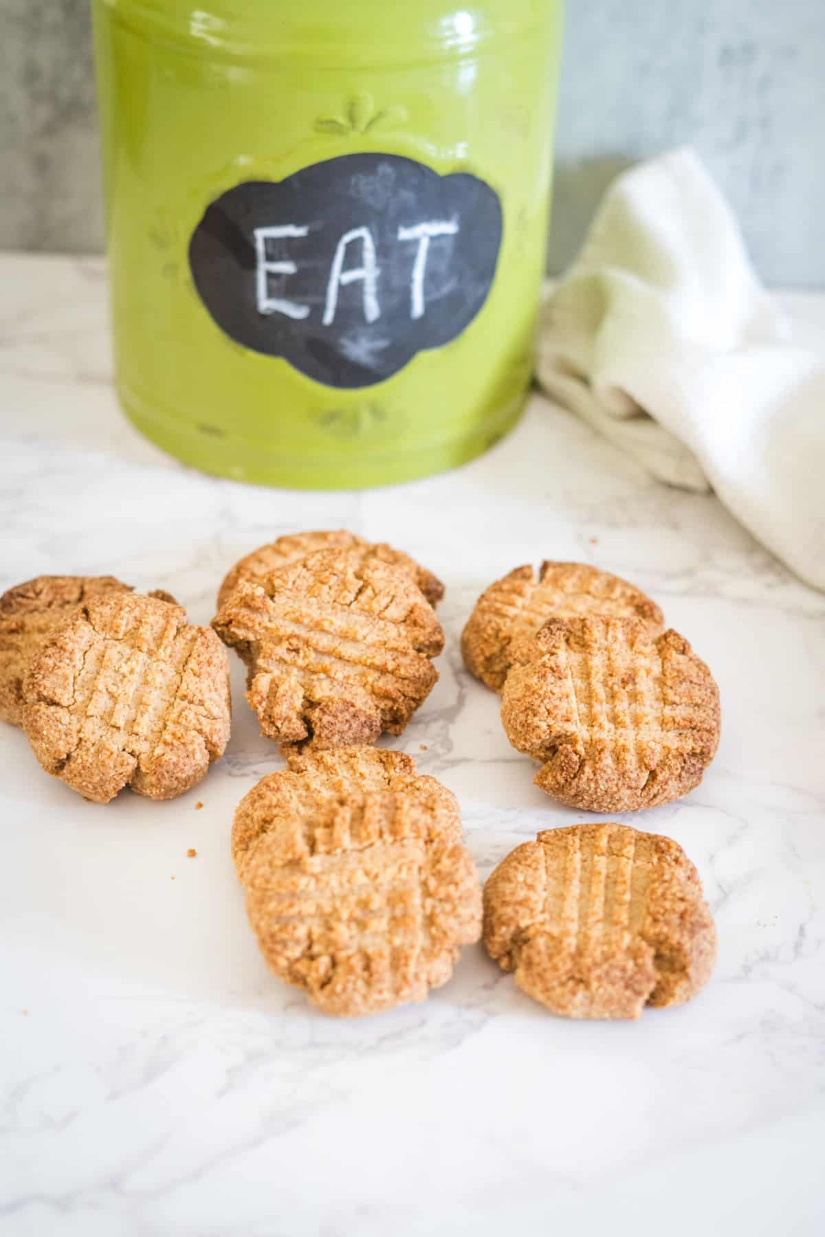 Six almond flour cookies are arranged on a marble surface in front of a green jar labeled "EAT." A white cloth is partially visible to the right.