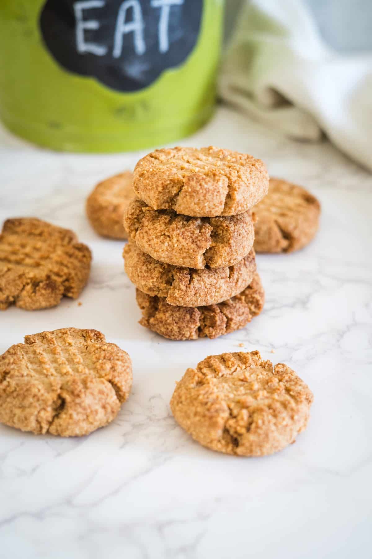 A stack of five peanut butter cookies and four additional cookies are placed on a marble surface with a green container labeled "EAT" in the background.