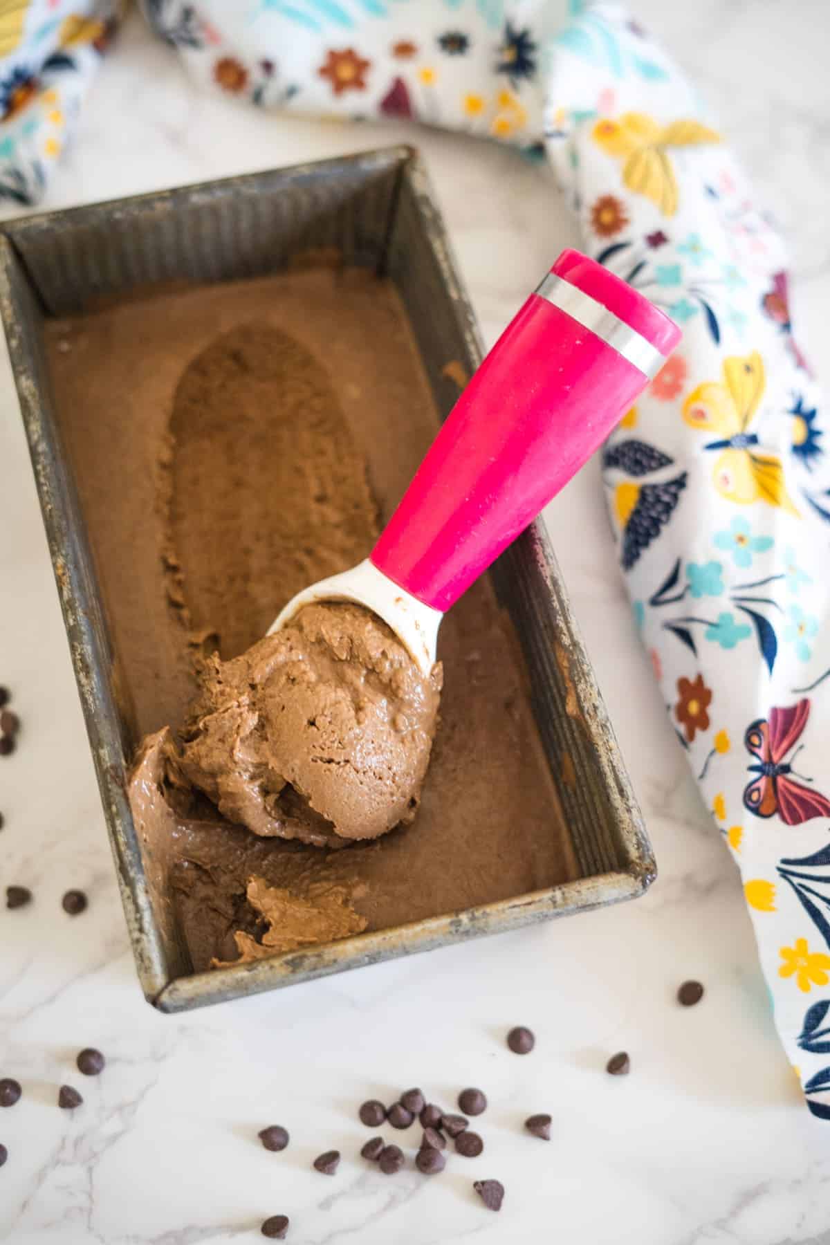 A pan of chocolate ice cream being scooped with a pink-handled scooper, surrounded by a colorful, butterfly-patterned cloth and scattered chocolate chips.