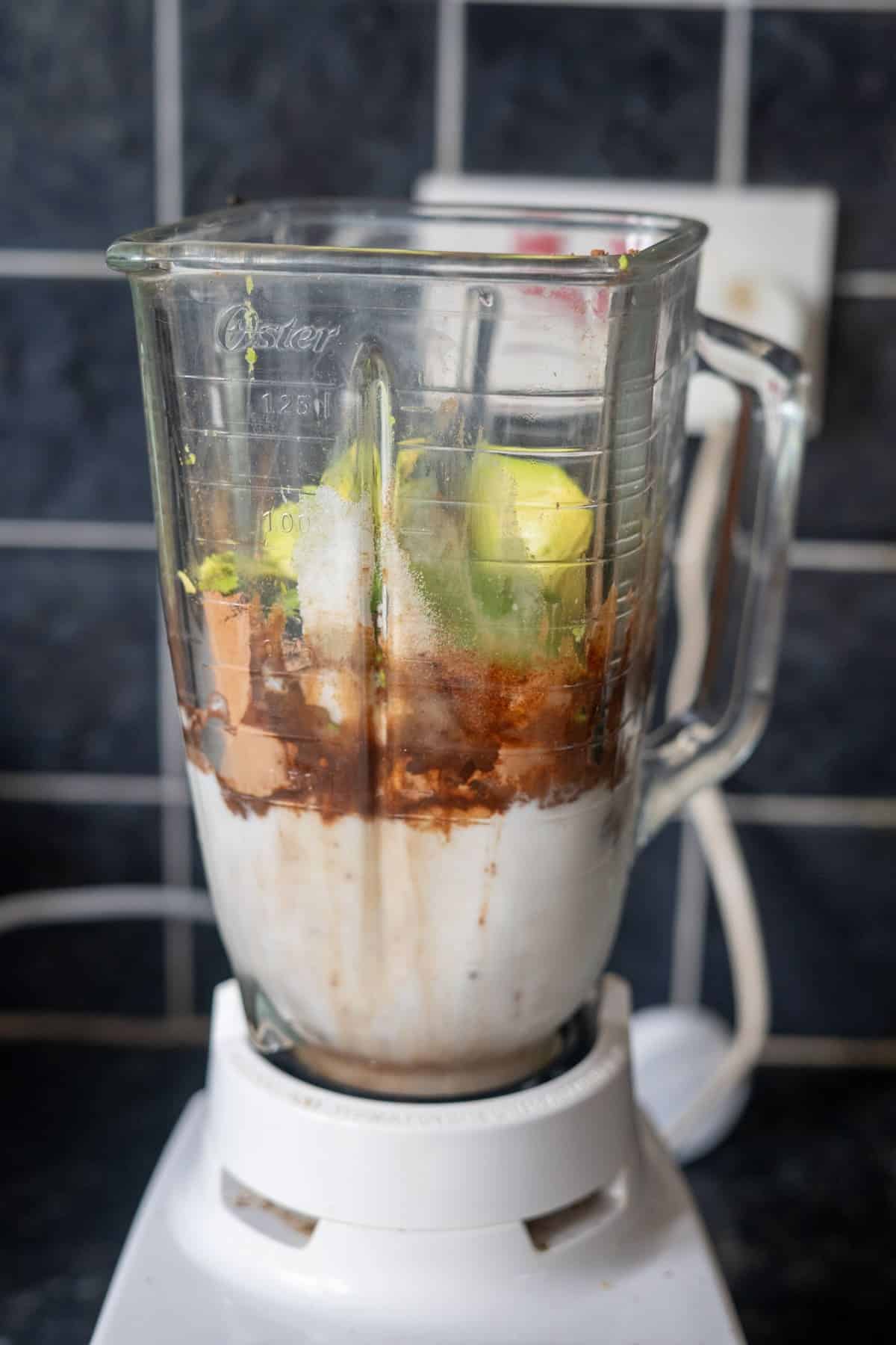 A blender jug filled with various ingredients, including avocado, cocoa powder, and a white liquid, stands on a white blender base in a kitchen.