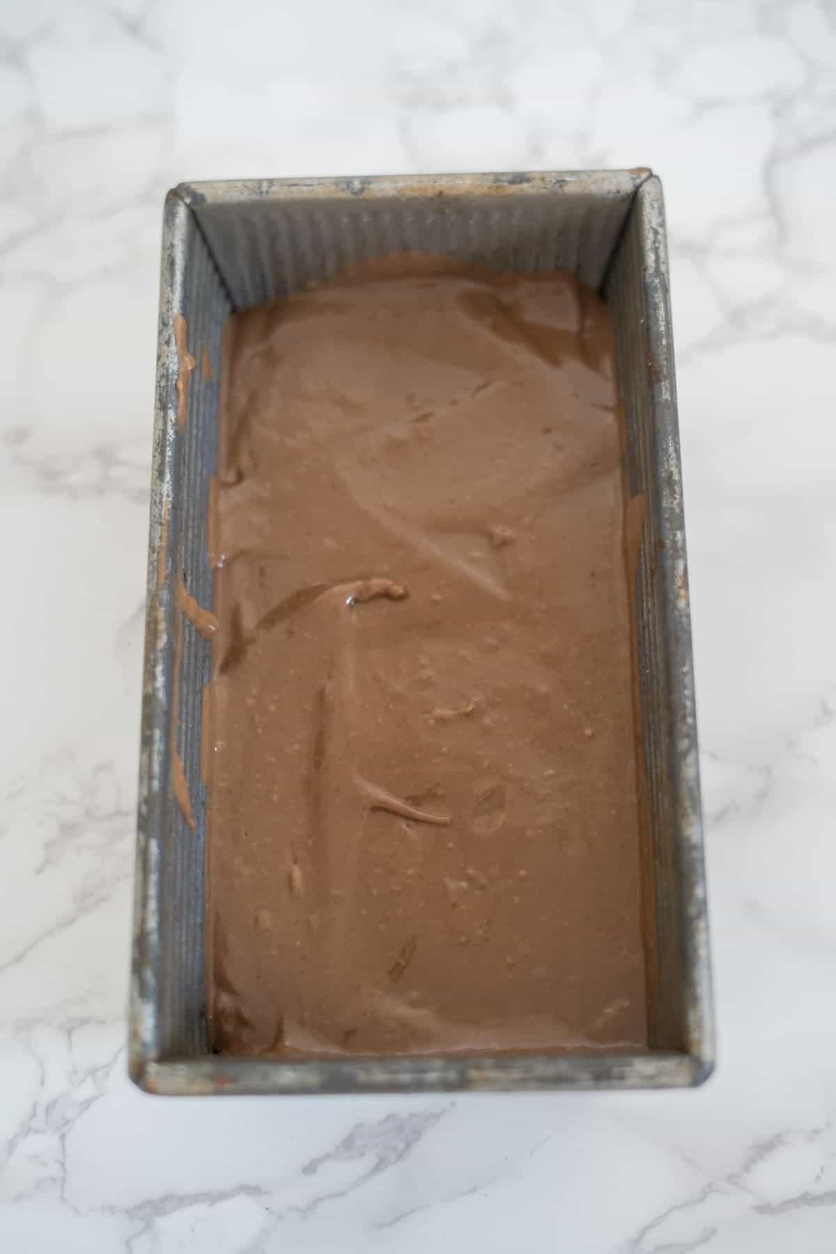 A rectangular metal loaf pan filled with chocolate batter, placed on a white marble surface.