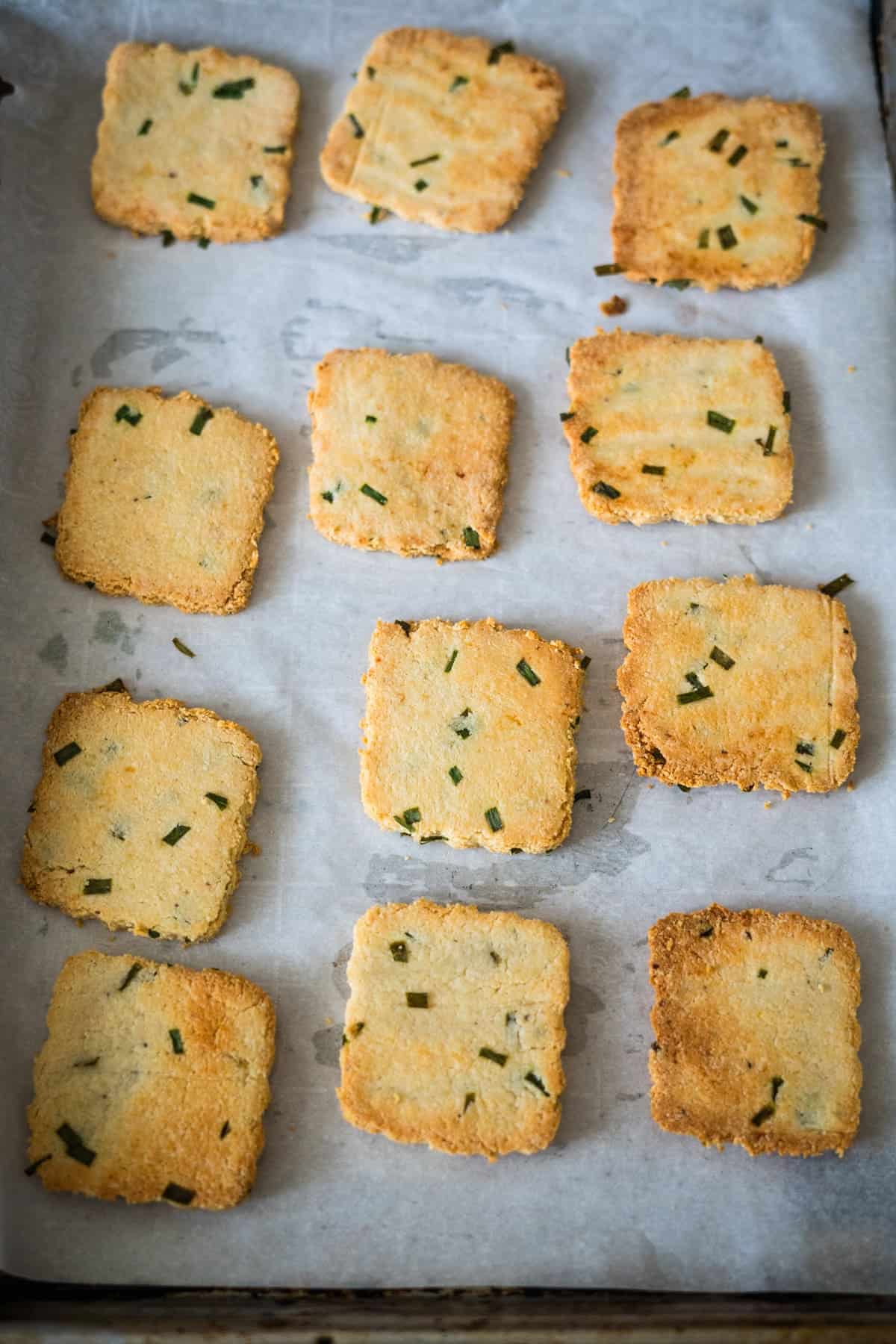 Twelve square, baked cookies with green herbs on a parchment-lined baking tray.