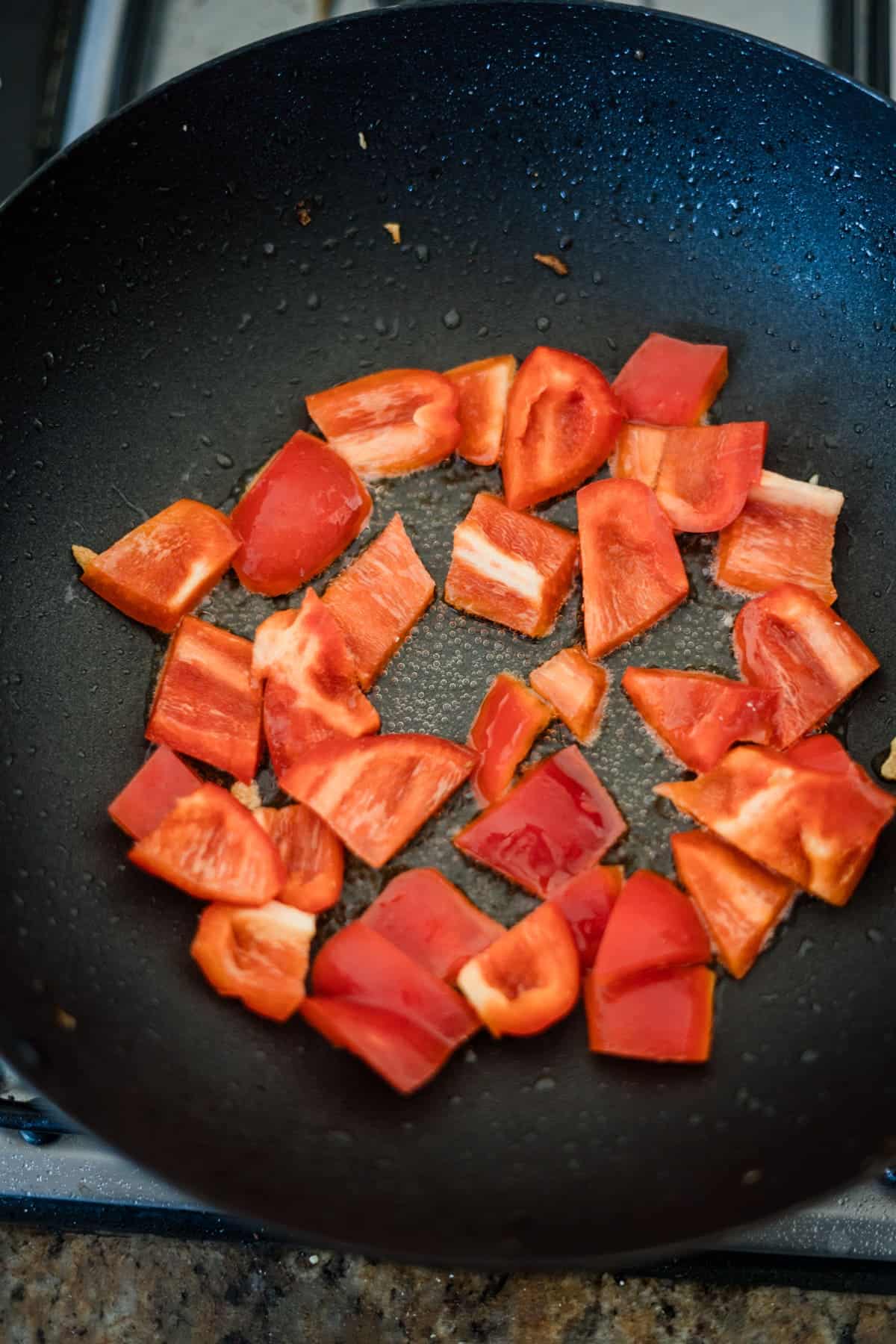 Chopped red bell pepper pieces and diced eggplant are being sautéed in a black frying pan on a stovetop, forming the base for a delicious tofu recipe.