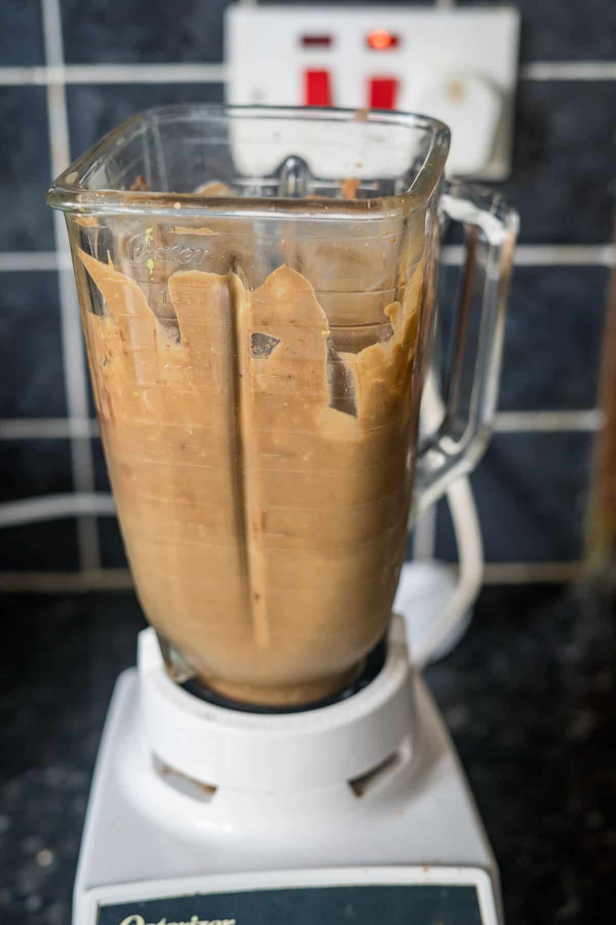 A blender filled with a thick brown mixture sits on a white base on a countertop, with a tiled backsplash and electrical outlet in the background.