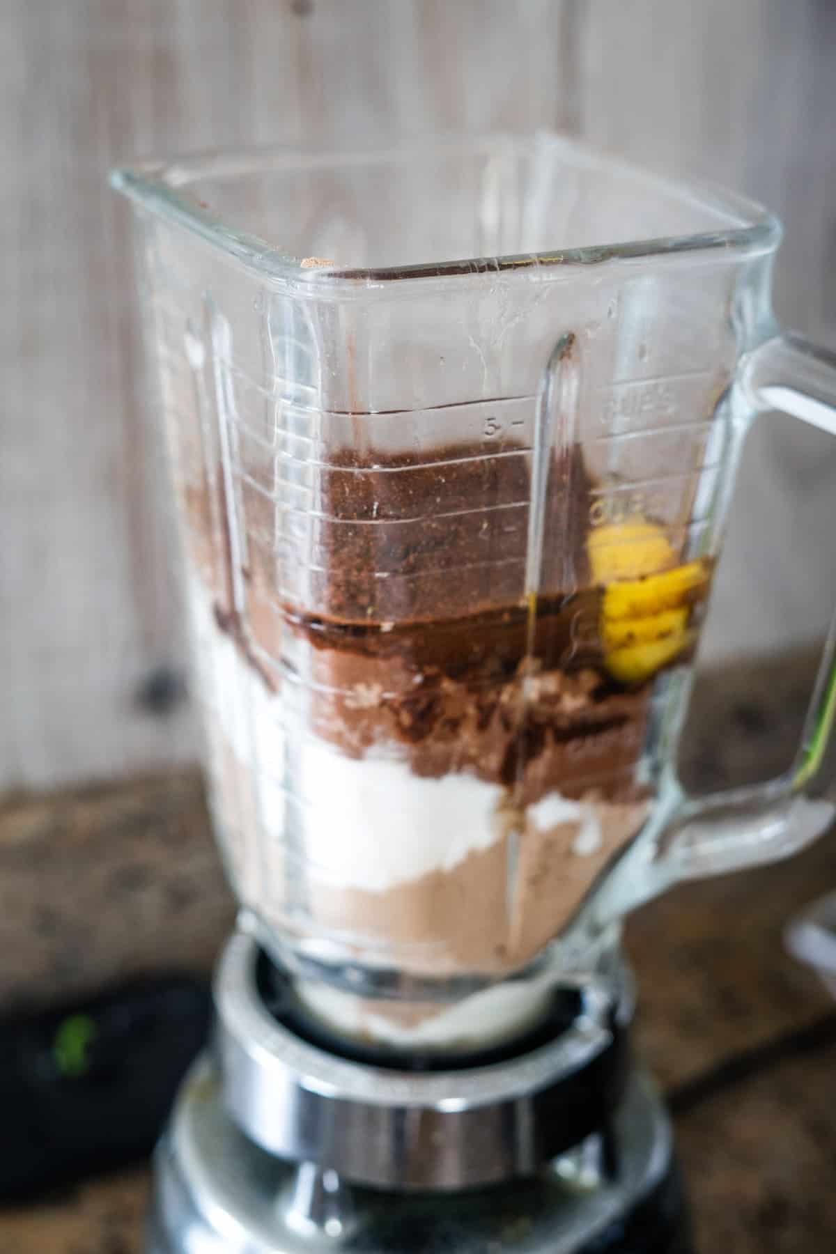 A blender containing measured ingredients, including powder, cocoa, bananas, and yogurt, sits on a wooden surface—all set to make delicious cottage cheese brownies.