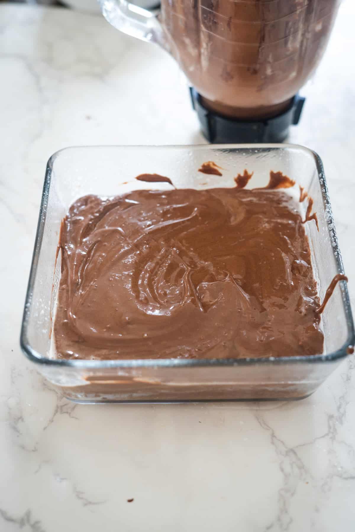 A glass container filled with a thick, chocolate mixture sits on a marble countertop, promising irresistible cottage cheese brownies. A blender with more of the mixture is visible in the background.