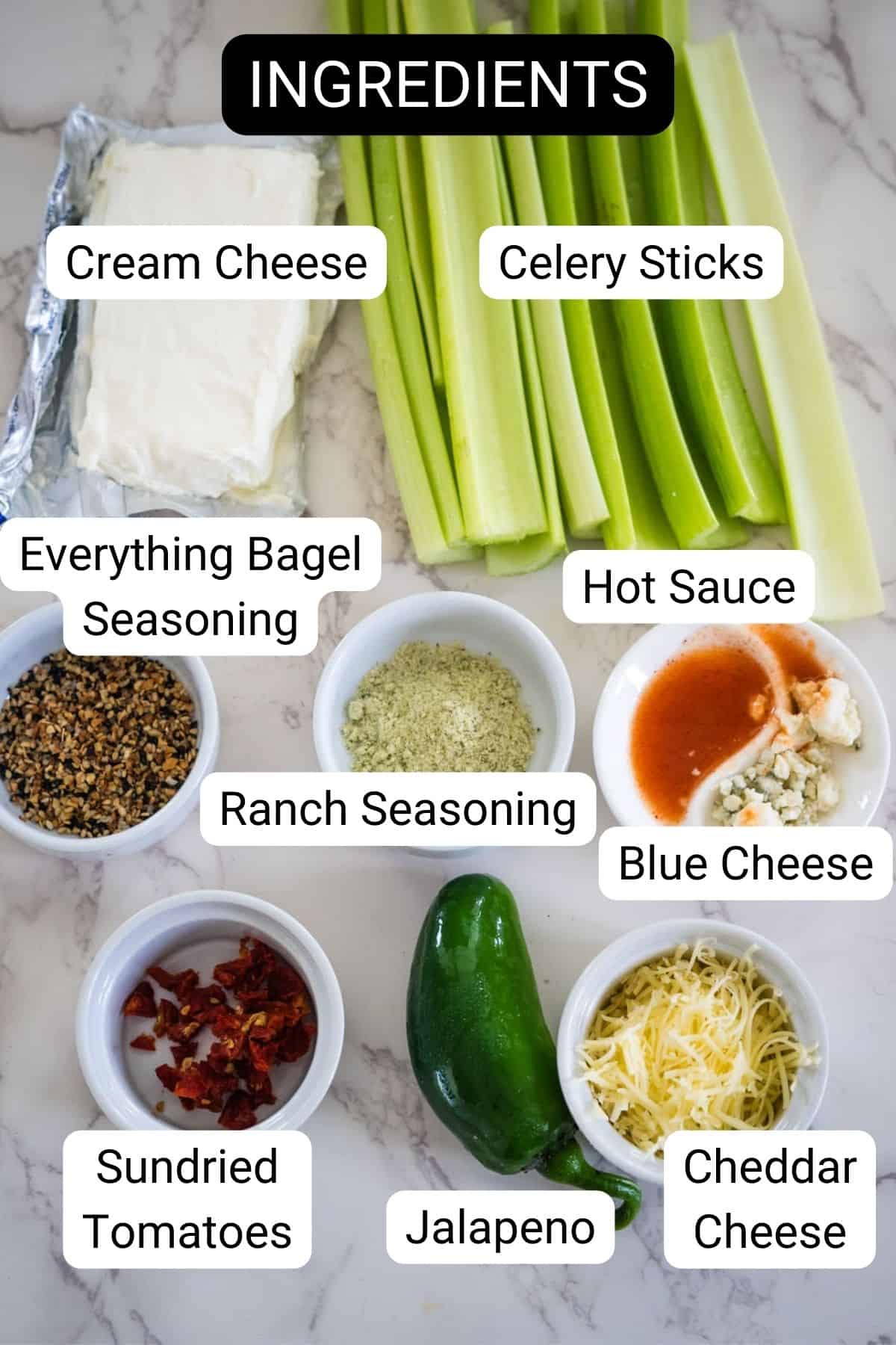 A display of ingredients including cream cheese, celery sticks, everything bagel seasoning, ranch seasoning, hot sauce, blue cheese, sundried tomatoes, jalapeno, and cheddar cheese on a marble surface.