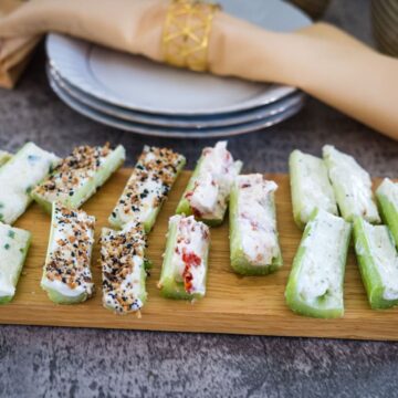 Celery sticks filled with various cream cheese spreads are arranged on a wooden board. Plates and a napkin with a gold napkin ring are in the background.