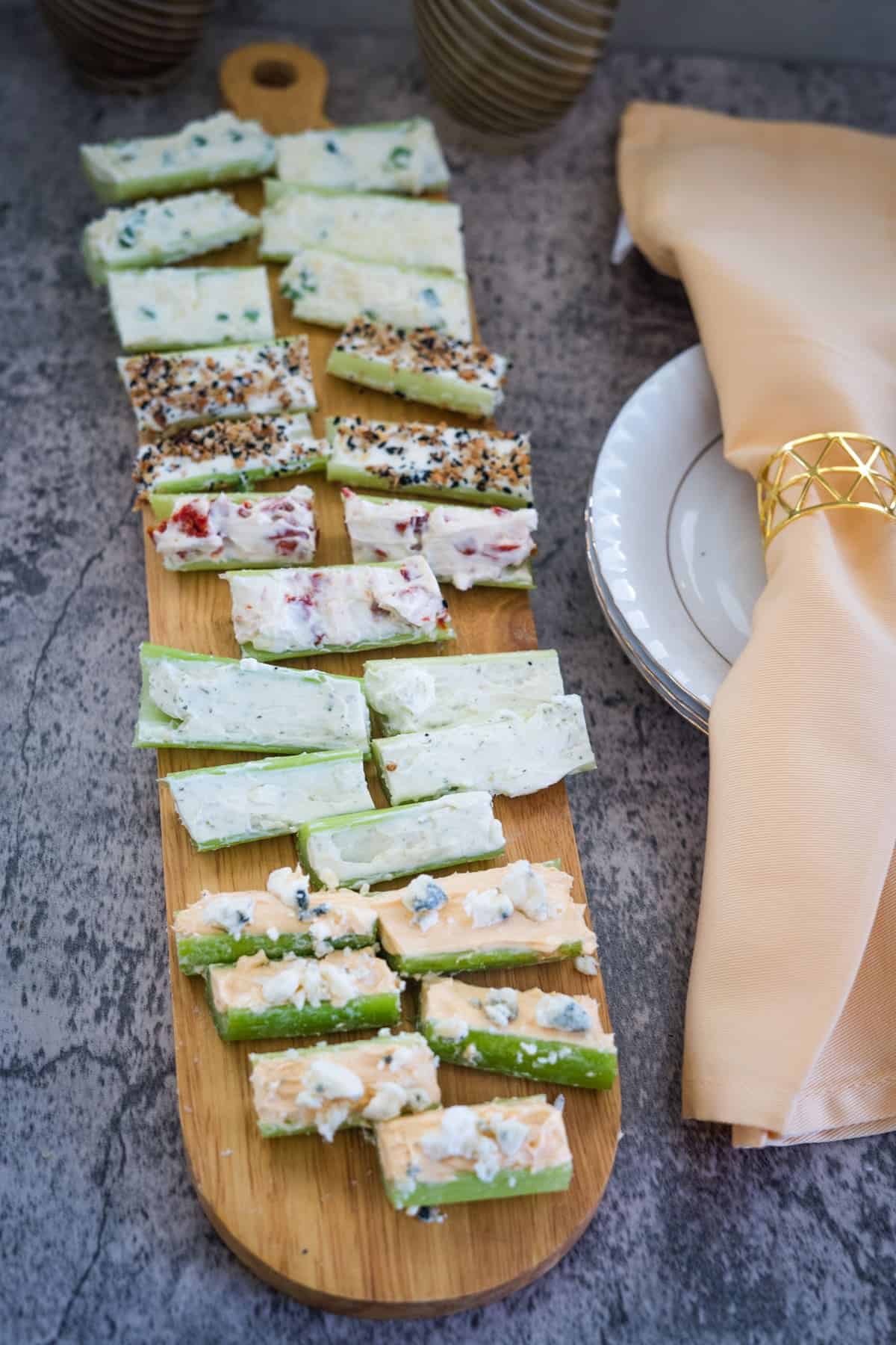 A wooden board holds rows of cucumber slices topped with various spreads and garnishes, next to a folded beige napkin with a gold napkin ring on a white plate.