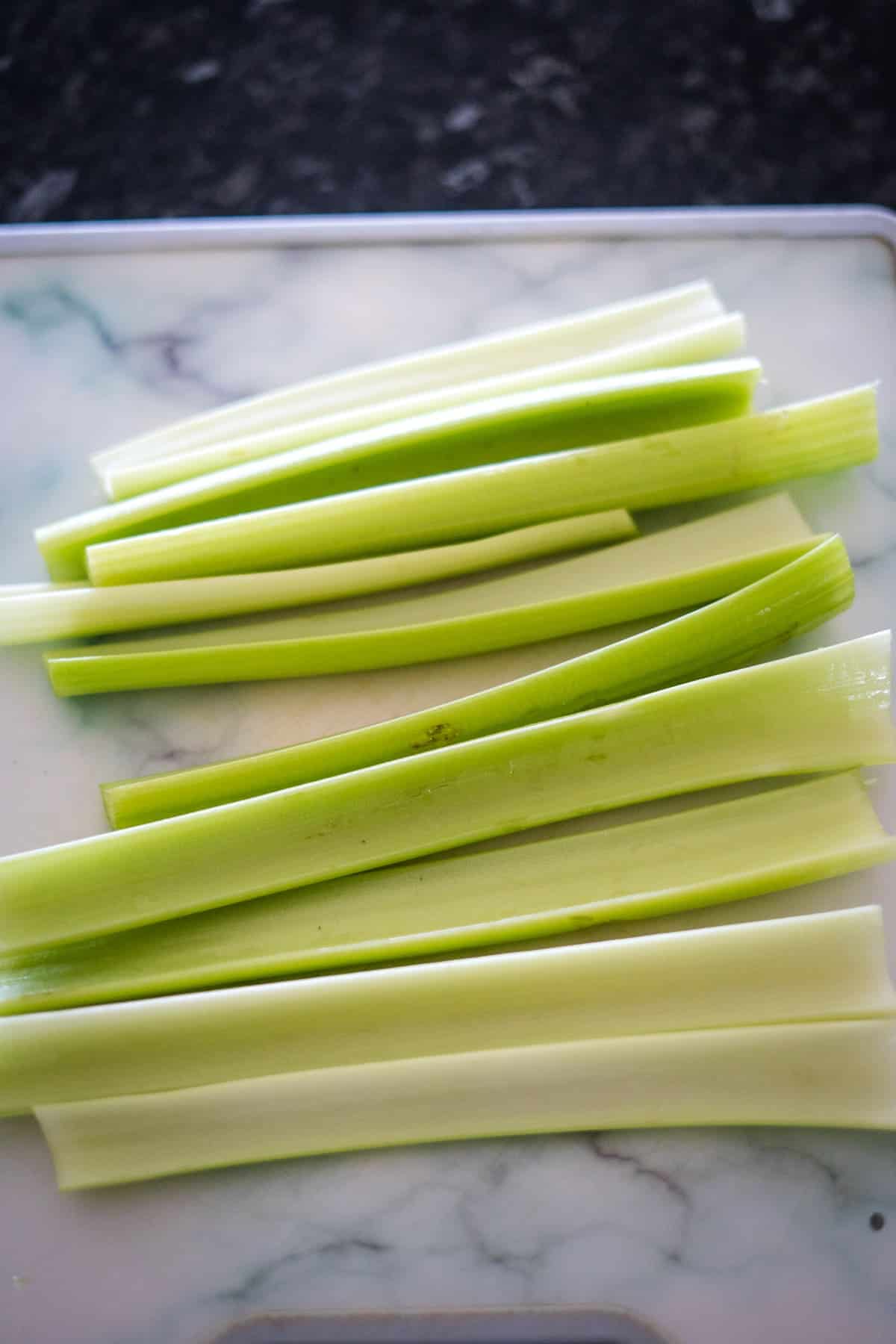 Several stalks of fresh green celery, perfect for cream cheese stuffed celery, are laid out on a white and marble cutting board.