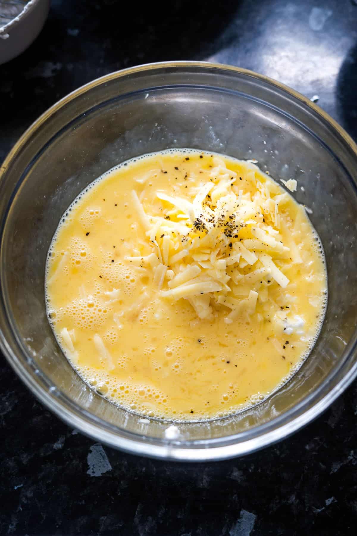 A glass bowl contains beaten eggs mixed with shredded cheese, cottage cheese, and pepper.