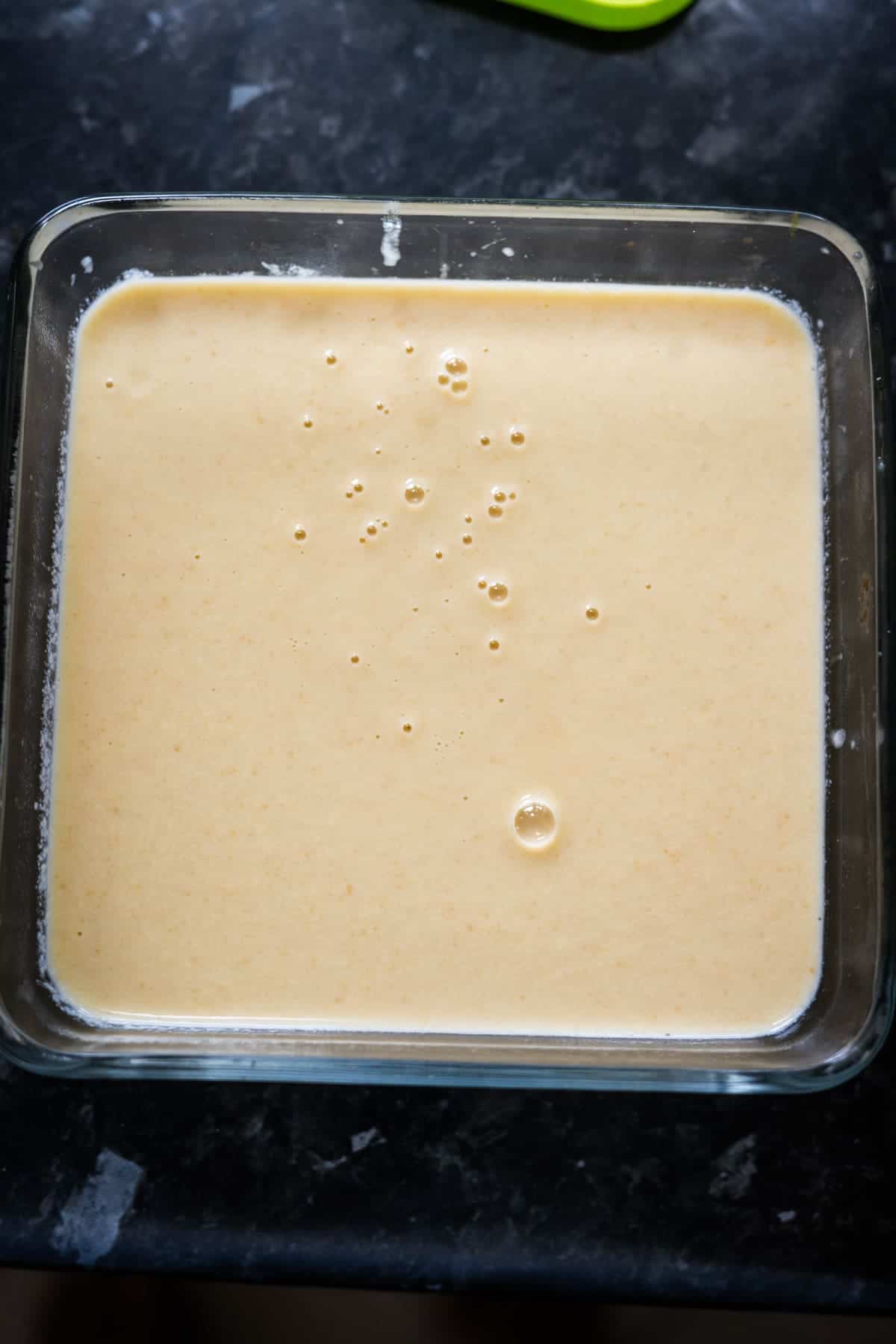 A square glass dish filled with a smooth, light-colored liquid mixture, possibly keto vanilla ice cream batter or custard, sits on a dark countertop.