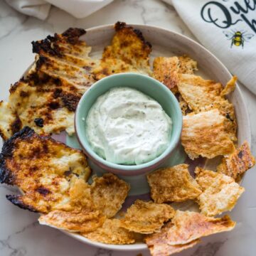 A plate of various toasted pita chips encircles a bowl of creamy white dip on a marble countertop.