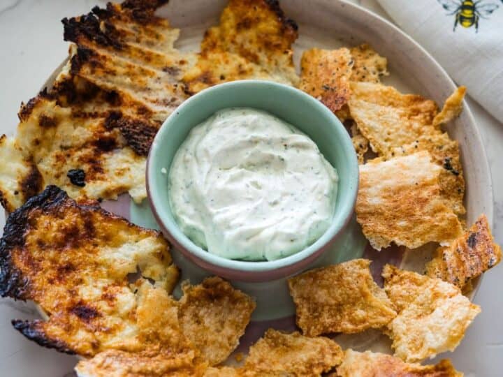 A plate of various toasted pita chips encircles a bowl of creamy white dip on a marble countertop.