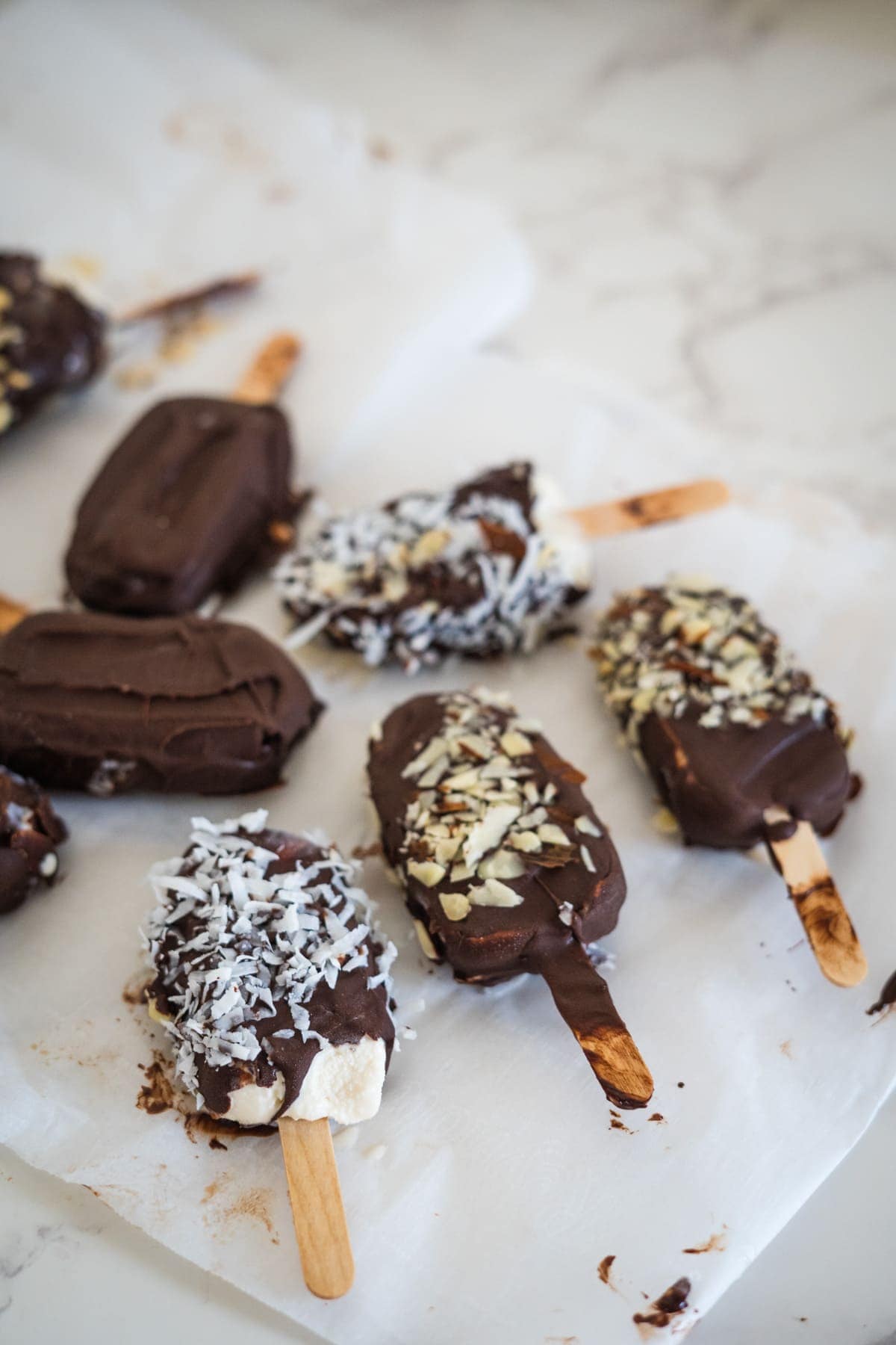 Keto ice cream bars coated in rich chocolate, topped with shredded coconut and chopped nuts, arranged on parchment paper.