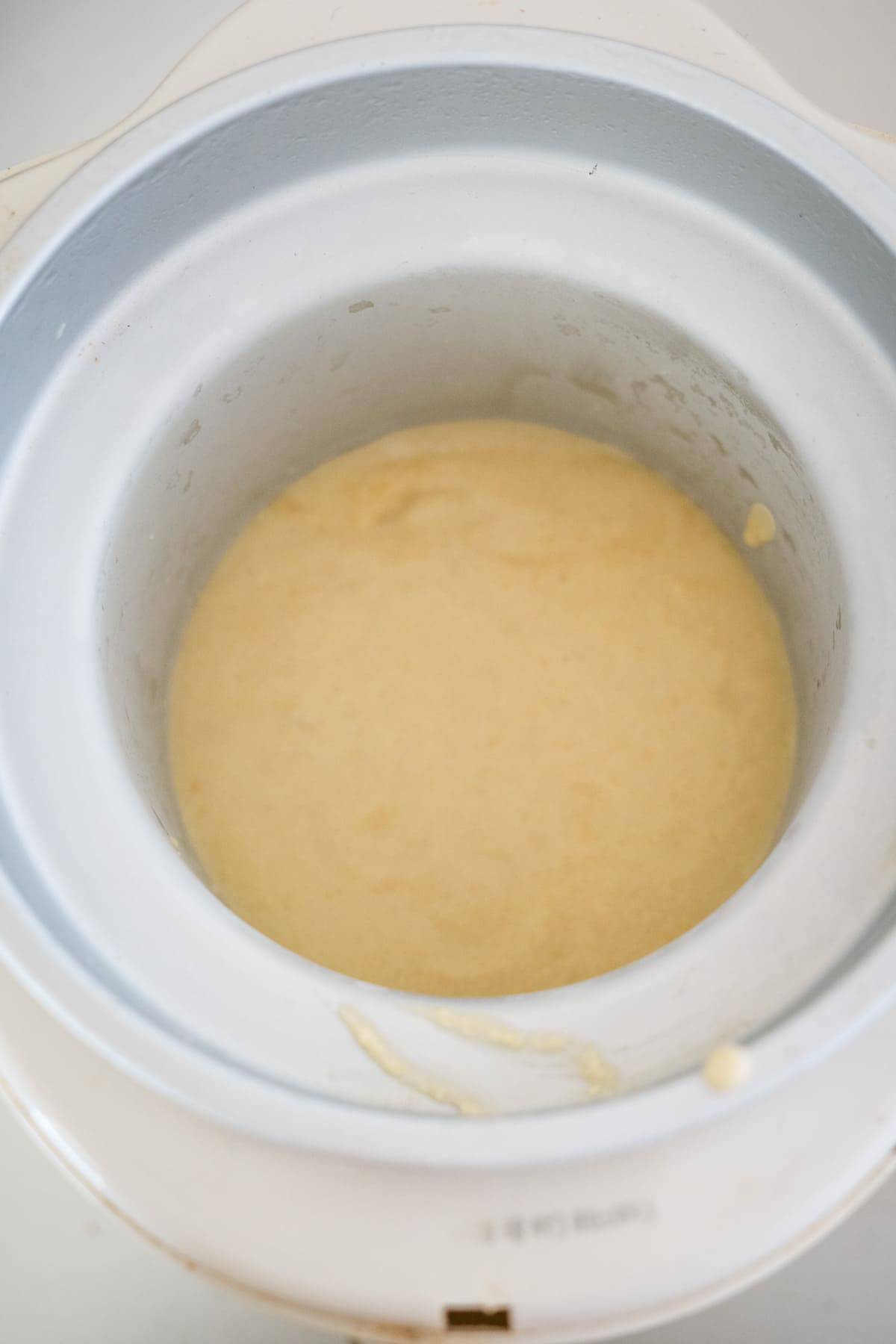 Top-down view of an electric rice cooker containing a mixture in the early stages of cooking. The mixture, which could be the beginnings of keto vanilla ice cream, appears to be a batter or dough. The cooker has a white exterior and a removable inner pot.