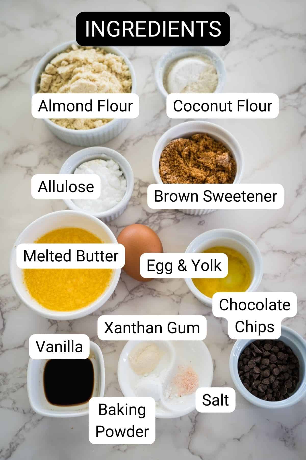 A display of various ingredients in bowls labeled as almond flour, coconut flour, allulose, brown sweetener, melted butter, egg & yolk, vanilla, xanthan gum, baking powder, salt, and chocolate chips.