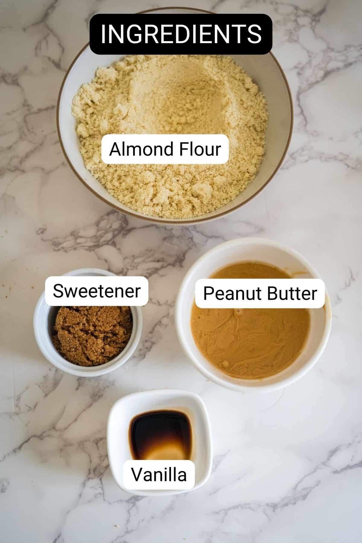 Overhead view of a bowl of almond flour, a small bowl of sweetener, a bowl of peanut butter, and a small dish of vanilla, labeled as ingredients, on a marble countertop.