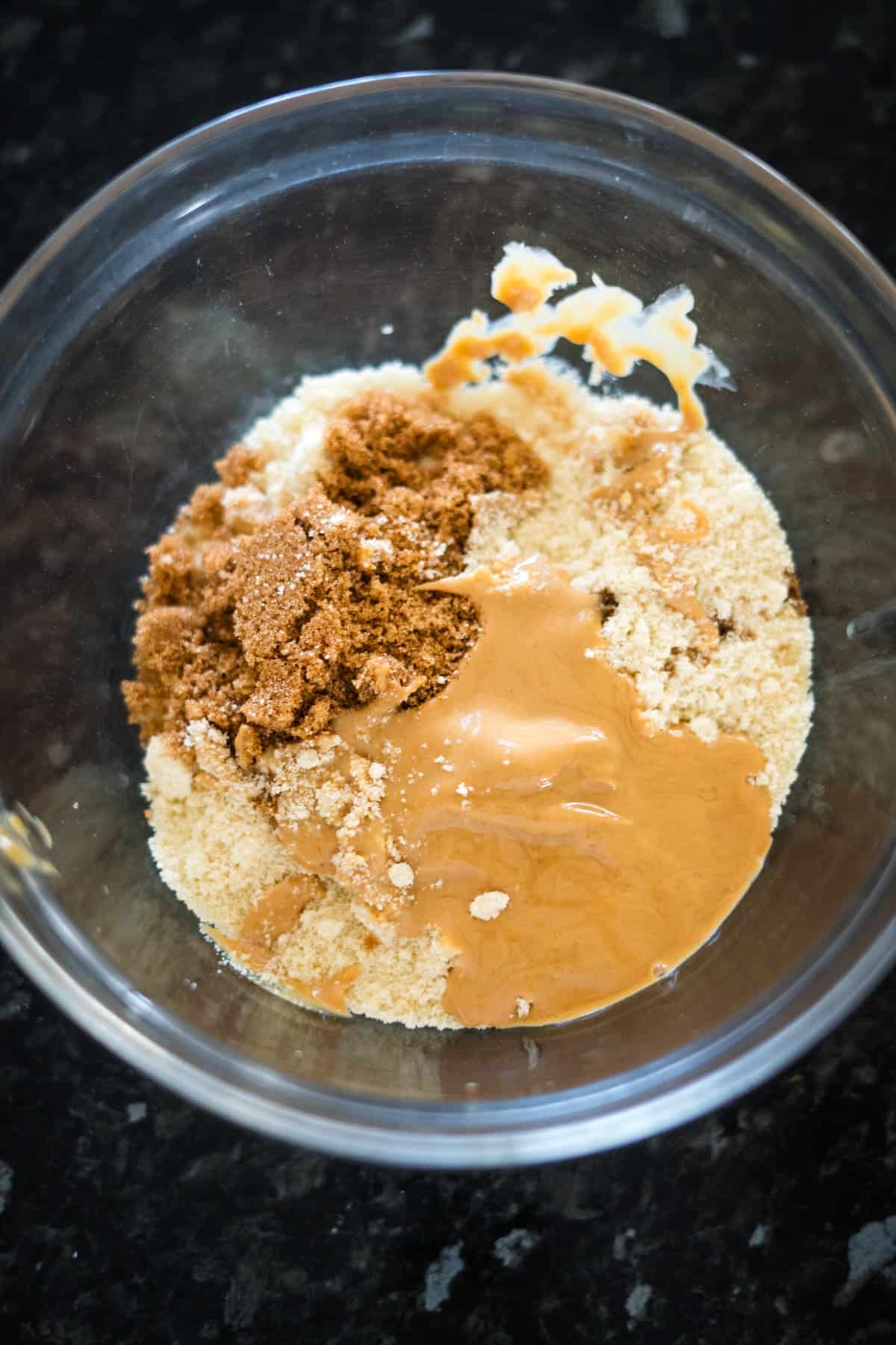 A glass bowl containing almond flour, brown sugar, and peanut butter on a dark countertop.