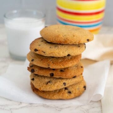 A stack of chocolate chip cookies on a white napkin with a glass of milk and a colorful striped mug in the background.