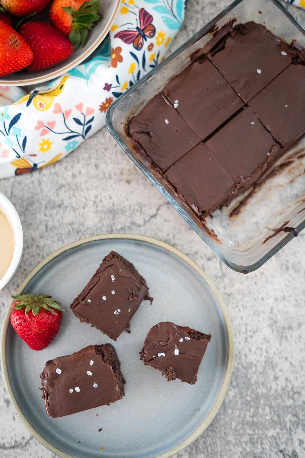 A plate with three squares of chocolate cottage cheese brownies topped with sea salt sits next to a glass dish containing six more squares. Fresh strawberries and a floral cloth can be seen in the background.