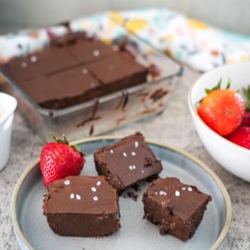 A plate of chocolate fudge squares with a sprinkling of sea salt next to whole strawberries, with additional fudge squares in a glass baking dish in the background.