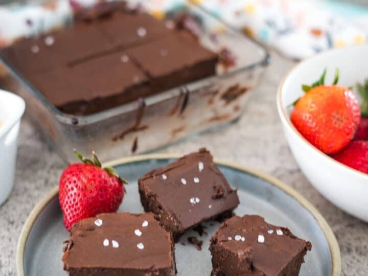 A plate of chocolate fudge squares with a sprinkling of sea salt next to whole strawberries, with additional fudge squares in a glass baking dish in the background.