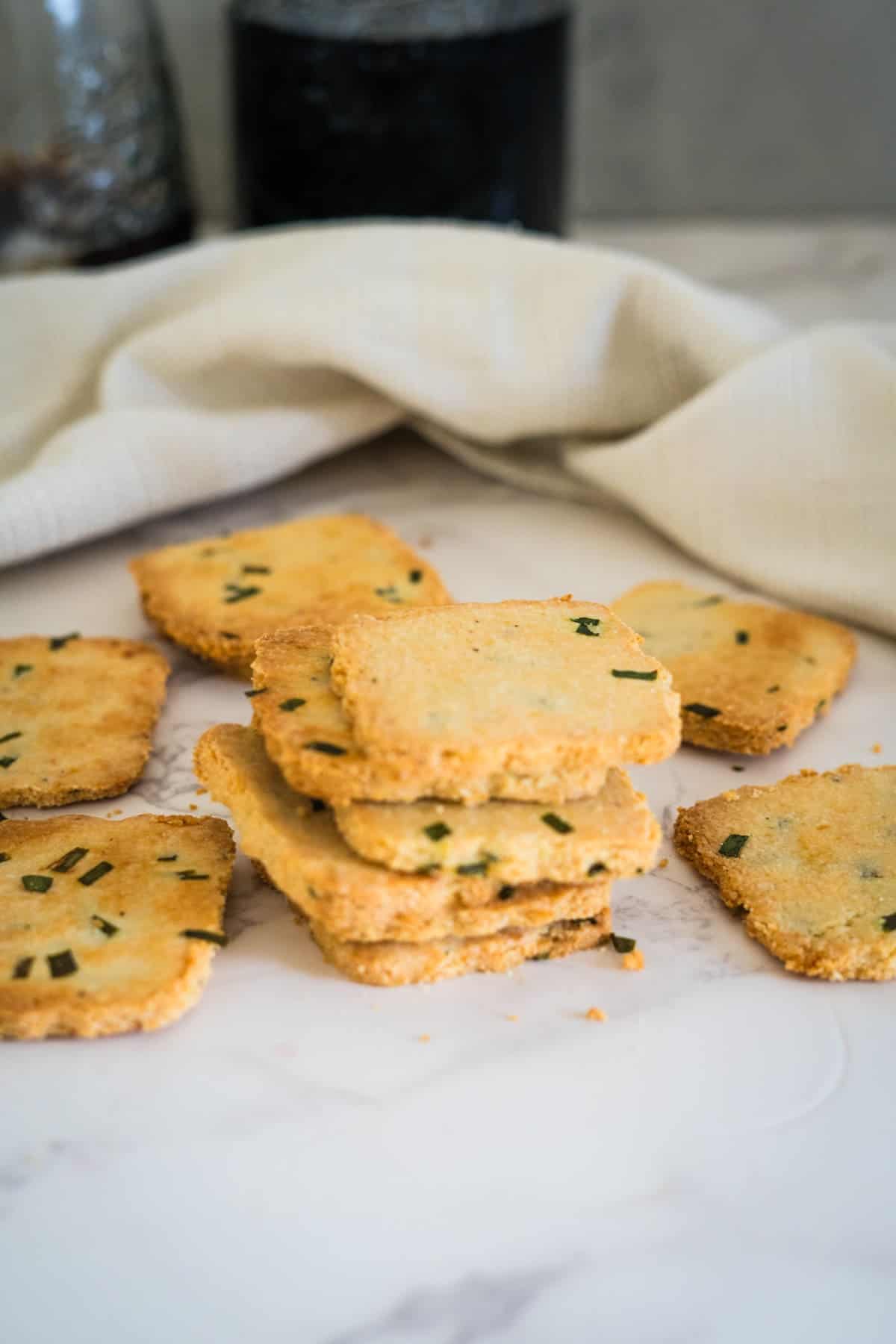 A stack of square, golden-brown cottage cheese crackers with green herbs are placed on a marble surface, with a white cloth in the background.