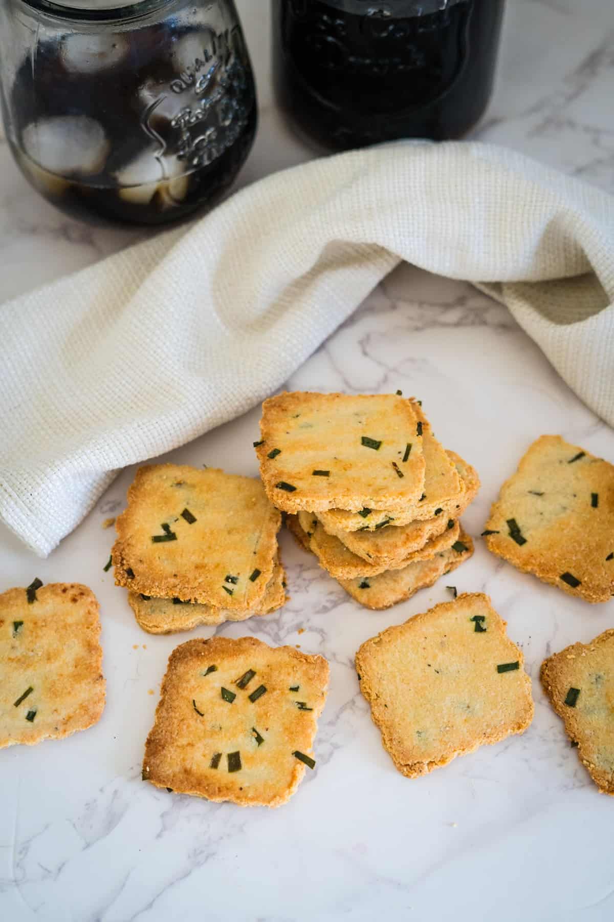 A stack of square, herb-topped crackers on a marble surface beside a white cloth and two jars filled with a dark beverage.