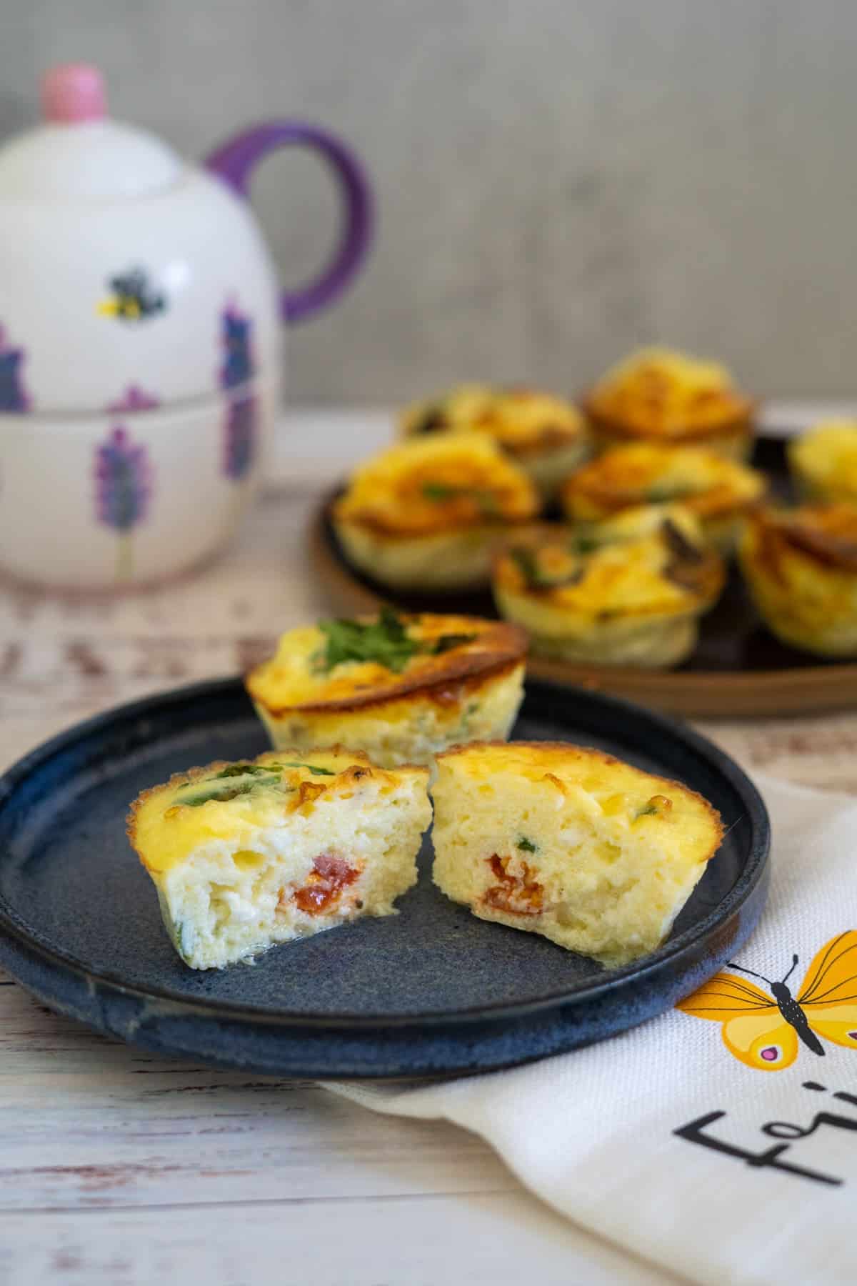 Close-up of two mini quiches on a dark plate, one cut in half to show the filling. A plate of more mini quiches in the background, with a teapot and a white napkin featuring a yellow butterfly.