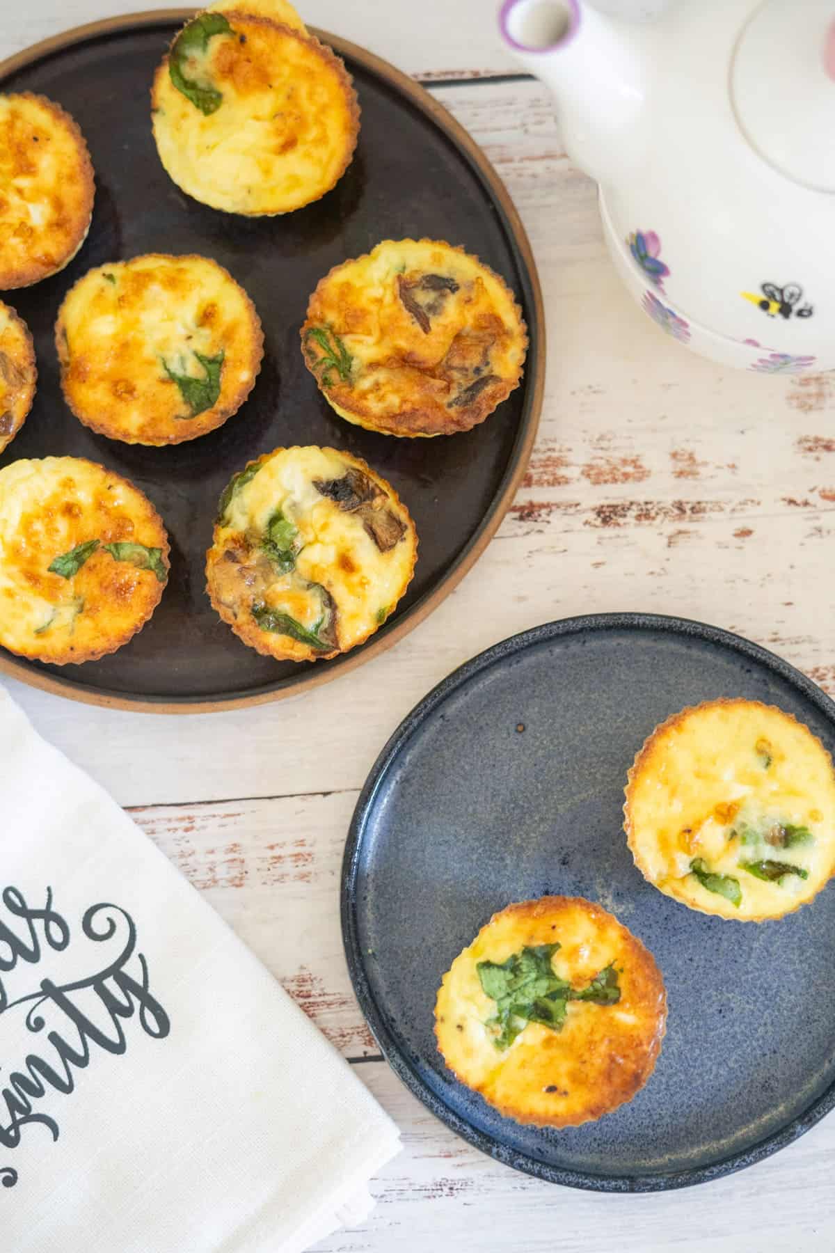 Plates with mini vegetable frittatas, cottage cheese egg muffins, a white tea towel, and a floral teapot rest on a white wooden table.